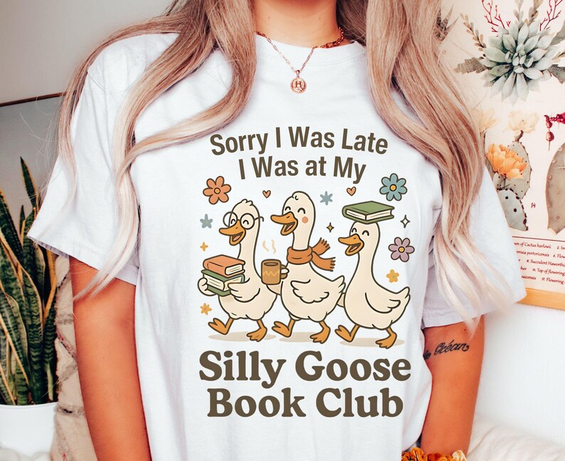 Pode incluir: Camiseta branca com o texto "Sorry I Was Late I Was at My Silly Goose Book Club". O design apresenta tr&ecirc;s gansos de desenho animado, cada um com livros ou uma x&iacute;cara de caf&eacute;, com motivos florais e estrelas.