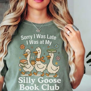 Pode incluir: T-shirt verde s&aacute;lvia com o texto "Sorry I Was Late, I Was at My Silly Goose Book Club". O gr&aacute;fico apresenta tr&ecirc;s gansos de desenho animado carregando livros e caf&eacute;. Flores e estrelas decorativas circundam os gansos.