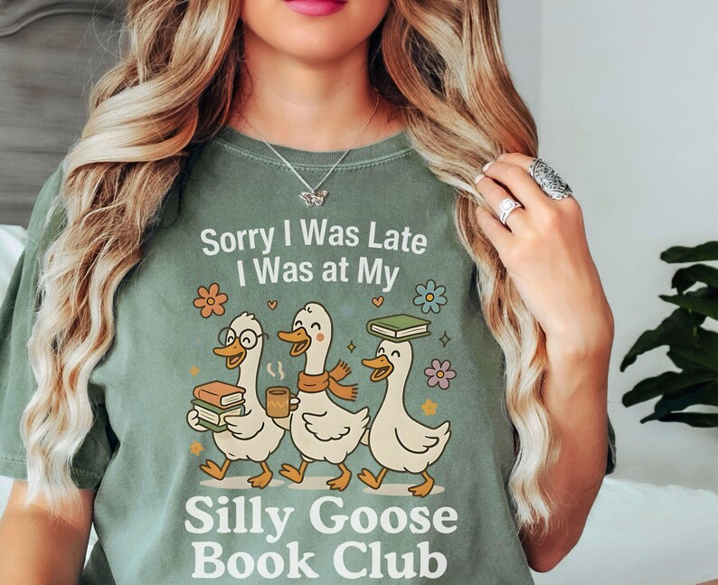 Pode incluir: T-shirt verde s&aacute;lvia com o texto "Sorry I Was Late, I Was at My Silly Goose Book Club". O gr&aacute;fico apresenta tr&ecirc;s gansos de desenho animado carregando livros e caf&eacute;. Flores e estrelas decorativas circundam os gansos.
