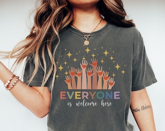 May include: A dark grey t-shirt with a graphic of raised hands with hearts and the text "EVERYONE is welcome here" in rainbow colours.