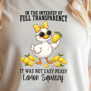 May include: White t-shirt featuring a cartoon duck wearing sunglasses and a bow, holding a lemon. Text on the shirt reads "IN THE INTEREST OF FULL TRANSPARENCY IT WAS NOT EASY PEASY Lemon Squeezy." The brand name "SUMMER BIRCH" is at the bottom.