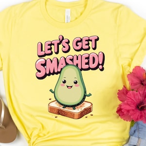Puede incluir: Camiseta amarilla con el texto "LET'S GET SMASHED!" y un aguacate de dibujos animados sobre una tostada. La imagen incluye pantalones cortos de mezclilla, gafas de sol y sandalias, sugiriendo un estilo casual.