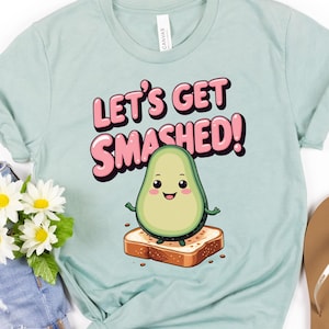 Puede incluir: Camiseta azul claro con el texto "Let's Get Smashed!" y un personaje de aguacate de dibujos animados sobre una tostada. La imagen también incluye pantalones cortos de mezclilla, flores blancas y amarillas y chanclas marrones.