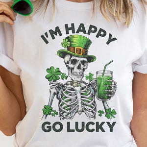 Op de afbeelding: Wit T-shirt met een St. Patrick's Day-afbeelding. Het ontwerp toont een skelet met een groene hoge hoed en een groen drankje. De tekst luidt "I'm Happy Go Lucky". Groene klavers zijn ook in het ontwerp opgenomen.