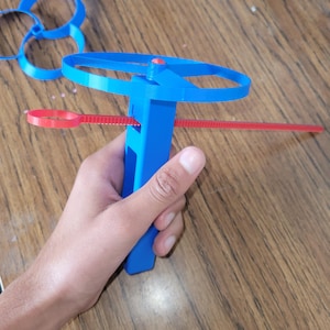 May include: A blue plastic toy with a red string attached to it. The toy has a propeller and a handle. The toy is being held by a hand.