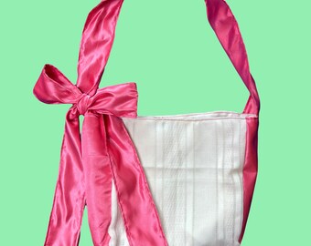 Peut inclure: Un sac cabas en tissu blanc avec un nœud en satin rose. Le sac a une bandoulière en satin rose.
