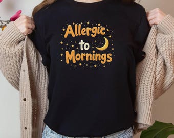 Puede incluir: Camiseta negra con el texto "Allergic to Mornings" en naranja, con un diseño de luna creciente y estrellas. La camiseta es de cuello redondo.