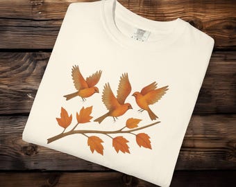 May include: Cream-colored t-shirt featuring a design of three orange birds with outstretched wings perched on a brown branch with orange leaves. The shirt is folded on a wooden surface.