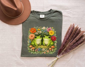 May include: Olive green t-shirt featuring a colorful illustration of two green frogs surrounded by flowers and mushrooms. A brown hat and dried pampas grass are also in the image. The shirt is a comfortable, casual style.