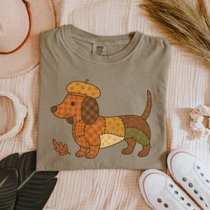 May include: A sage green t-shirt featuring a cartoon dachshund wearing a beret. The dog is made of patchwork fabric in shades of brown, orange, and green. A fall leaf is next to the dog. The shirt is displayed with a hat, scrunchie, and white sneakers.