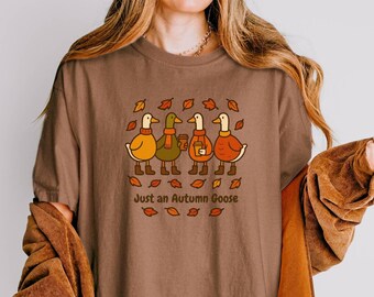 May include: Brown t-shirt featuring a graphic of four cartoon geese in fall attire, holding coffee cups and surrounded by leaves. The text "Just an Autumn Goose" is printed below the geese.