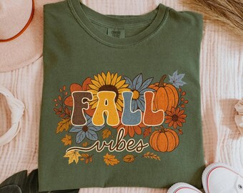 May include: Green t-shirt with a retro design featuring the words "FALL vibes" surrounded by sunflowers, pumpkins, and fall leaves. The shirt is a crew neck and is displayed on a textured surface.