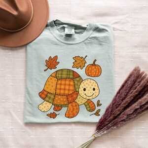 May include: A light blue t-shirt featuring a cartoon turtle with a patchwork shell design in fall colors. The turtle is surrounded by autumn leaves and a pumpkin. A brown hat is in the upper left corner.