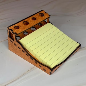 May include: A light brown wooden desk organizer with a slanted surface holding a stack of pale yellow lined sticky notes. The organizer features a top section with five circular cutouts for pens or pencils and has visible laser-cut joinery details.