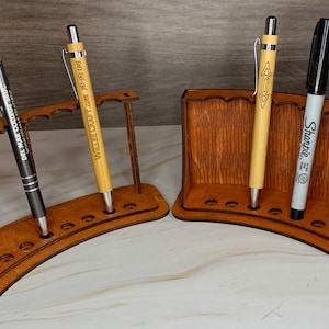 Könnte beinhalten: Zwei geschwungene Stiftehalter aus Holz mit mehreren runden Aussparungen für Schreibgeräte. Der linke Halter hat ein offenes Rahmendesign, während der rechte Halter eine solide Rückwand besitzt. Stifte und Marker stecken in den Öffnungen.