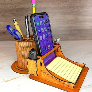 May include: A wooden desk organiser featuring a cylindrical pen holder with multiple slots, a central smartphone stand, and a small compartment for a stack of yellow lined sticky notes. The organiser is crafted from light brown wood with visible grain.