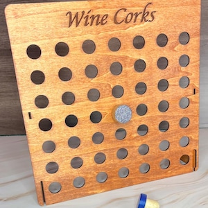 Puede incluir: Soporte de corchos de vino de madera con las palabras "Wine Corks" grabadas en la parte superior. El soporte tiene múltiples agujeros circulares para guardar corchos, con un corcho decorativo en el centro y un corcho de vino en la parte inferior.