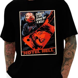 May include: Black t-shirt featuring a horror movie poster design. The poster has a red and orange color scheme with the text "CHECKING IN IS EASY... CHECKING OUT IS HELL!" and "MOTEL HELL". The design includes a pig mask and a cleaver.