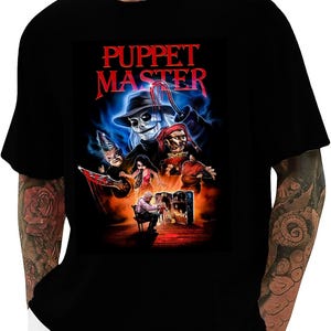 May include: Black t-shirt featuring the text "PUPPET MASTER" in red, above a graphic of several puppet characters. The graphic has a dark, horror movie theme with a blue and orange color scheme.