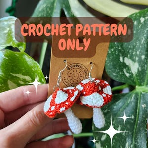Könnte beinhalten: Gehäkelte Pilzohrringe mit roten Kappen mit weißen Punkten und weißen Stielen. Im Hintergrund sind grüne Blätter zu sehen. Der Text "CROCHET PATTERN ONLY" ist im Bild enthalten.