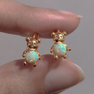 Puede incluir: Un par de pendientes de oro en forma de oso con piedras de ópalo. Los pendientes son pequeños y delicados.