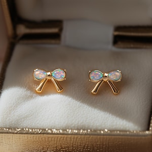 Könnte beinhalten: Ein Paar goldfarbene Ohrstecker in Schleifenform mit schillernden, opalartigen Zentren. Die Ohrringe werden in einer goldfarbenen Schmuckschatulle mit weißem Innenraum präsentiert. Ein zartes und feminines Accessoire.