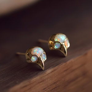 Peut inclure: Une paire de boucles d'oreilles à clous dorées. Chaque boucle d'oreille a la forme d'un crâne d'oiseau avec des pierres opalescentes incrustées dans les orbites et la tête. Les boucles d'oreilles sont présentées sur une surface en bois.