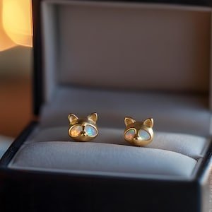 May include: A pair of gold-colored raccoon-shaped stud earrings with opal-like centers, displayed in a black jewelry box with a light-colored interior. The earrings have a polished finish and are set against a blurred background of warm, out-of-focus lights.