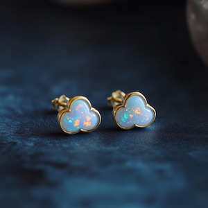 May include: A pair of cloud-shaped stud earrings. Each earring features a light blue opal center with gold-colored trim. The opals have hints of orange and green. The earrings are displayed on a dark blue surface.
