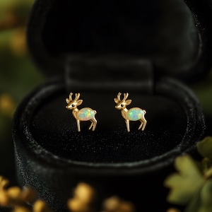 Puede incluir: Un par de pendientes de botón dorados con forma de ciervo y centros con aspecto de ópalo, presentados en una caja de joyería negra. Los pendientes tienen cuernos detallados y están de pie. El fondo está borroso con tonos verdes y amarillos.