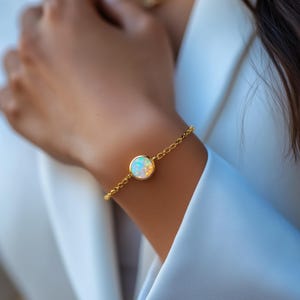 Puede incluir: Una pulsera dorada con una piedra de ópalo redonda. El ópalo muestra una mezcla de colores, incluyendo blanco, azul y amarillo. La pulsera es delicada, con un diseño de eslabones de cadena. La joya se lleva en la muñeca, sobre un fondo blanco.