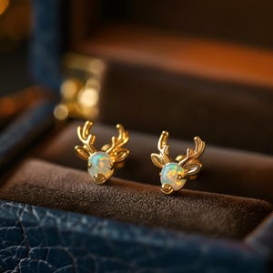 Puede incluir: Un par de pendientes dorados con forma de reno con piedras preciosas tipo ópalo. Los pendientes se muestran dentro de un joyero azul oscuro con interior marrón. Los pendientes tienen un diseño detallado de cuernos.