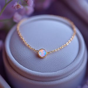 以下が含まれることがあります： 丸いオパール宝石をあしらった繊細なゴールドブレスレット。 オパールはパステルカラーのミックスで、ゴールドベゼルにセットされています。 ブレスレットは、背景に柔らかな紫色の花を添えて、灰色のジュエリーボックスに展示されています。