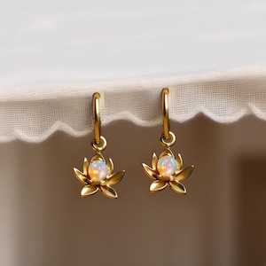 Könnte beinhalten: Ein Paar goldfarbene Creolen. Jede Ohrring hat ein Lotusblumen-Design mit einem zentralen opalartigen Stein. Die Ohrringe werden vor einem weißen, strukturierten Hintergrund präsentiert.