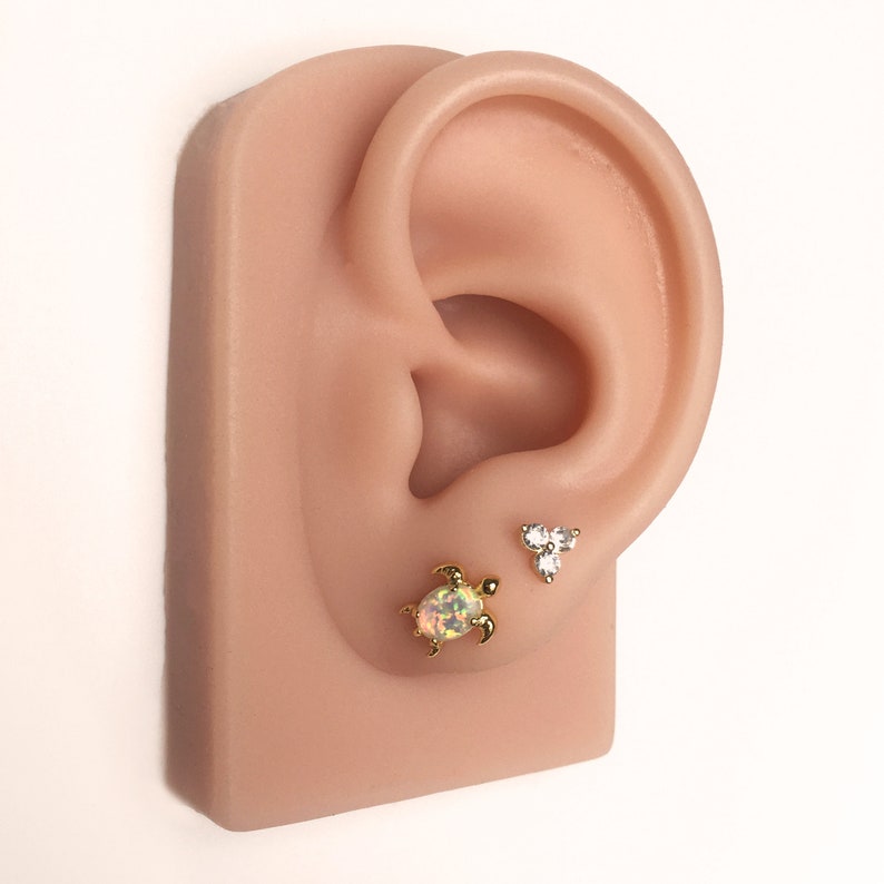 May include: A pair of gold earrings featuring a small turtle charm and a three-stone cluster design. The turtle charm has an iridescent opal stone in the centre.
