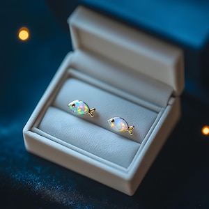 May include: Gold-coloured fish-shaped stud earrings with iridescent opal-like centres, displayed in an open, white jewellery box with a light blue interior. The earrings feature small, detailed fins.