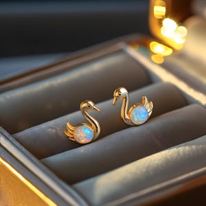 May include: A pair of gold-colored swan stud earrings. Each swan features a pearlescent, opalescent stone in the center of its body. The earrings are displayed in a gray jewelry box, with soft lighting highlighting the details.