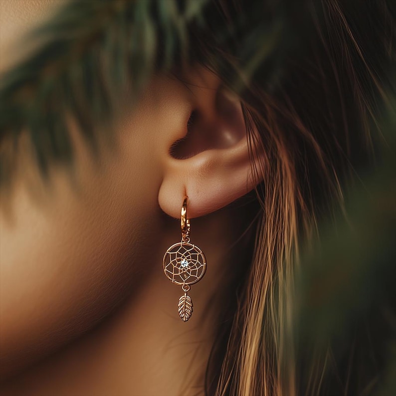 May include: Gold-colored dreamcatcher earring with a small diamond-like accent in the center and a dangling feather charm. The earring is attached to a gold hoop. The earring is worn on the ear of a person.