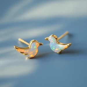 May include: A pair of gold-coloured bird-shaped stud earrings. Each earring has a white opal body with iridescent colour flashes and gold-coloured accents. The earrings are set against a blue background.