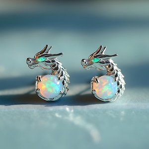 Puede incluir: Un par de pendientes de botón plateados con forma de cabeza de dragón, cada uno con un pequeño ojo verde brillante y una piedra redonda blanca iridiscente similar a un ópalo, sostenida por la cola curvada y la pata delantera con garras.