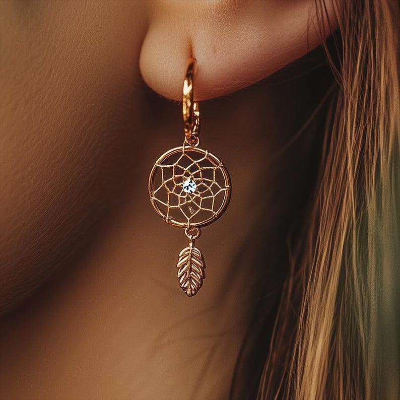May include: Gold-toned hoop earring featuring a dreamcatcher design with a central gemstone and a dangling feather charm. The intricate web-like pattern and delicate feather create a bohemian aesthetic. The earring is shown close-up against a blurred background.