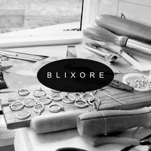 May include: A workbench with jewelry-making tools and materials. The word "BLIXORE" is displayed on a black oval. Silver rings are arranged on a wooden board, alongside pliers, a mandrel, and other tools.