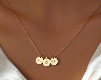 May include: A gold-colored necklace with three circular pendants. The pendants have the names "Ella" and "Sebi" in cursive script, and a paw print. The necklace is worn against a white shirt.