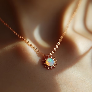 Op de afbeelding: Een delicate gouden ketting met een zonnehanger en een centrale opaalsteen. Het sunburst-ontwerp heeft puntige stralen rond de iriserende opaal. De ketting is een fijne schakelstijl en de ketting wordt getoond tegen een vage achtergrond.