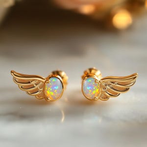 May include: A pair of gold-colored stud earrings. Each earring features an oval, iridescent opal stone set within a gold wing design. The wings have detailed feathering. The earrings are displayed on a light-colored surface.