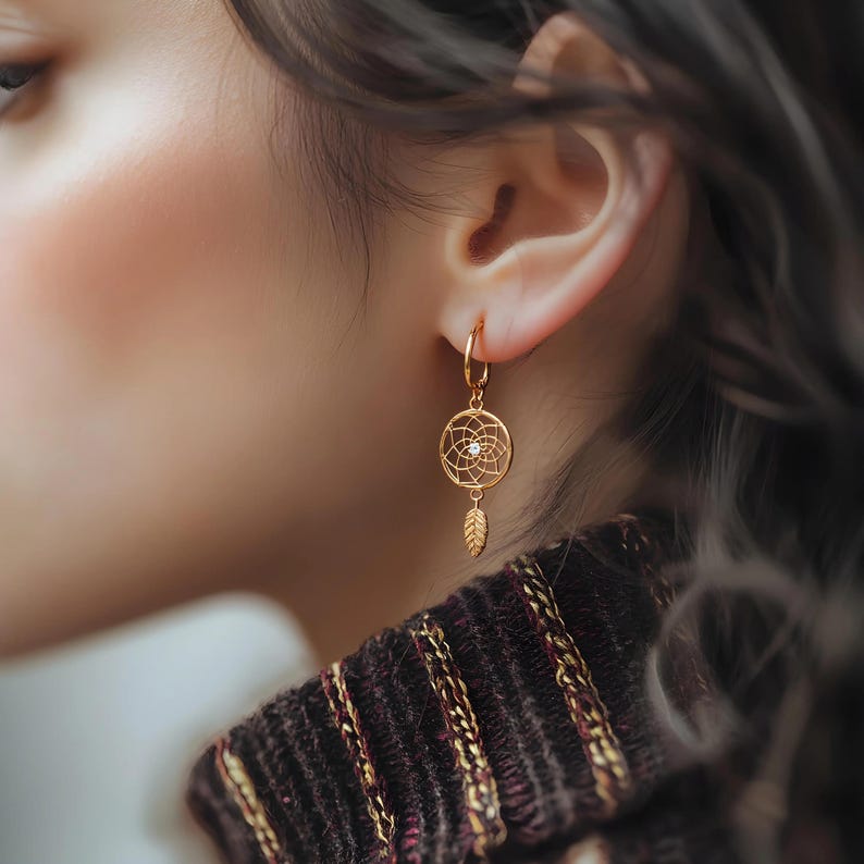 May include: Gold-colored hoop earring featuring a dreamcatcher design with a central clear stone and a dangling feather. The earring is worn on the ear, showcasing its intricate details and warm metallic hue.