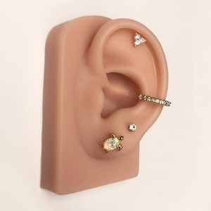 May include: A close-up of a fake ear with multiple piercings. The ear has a small gold turtle stud with an opal stone, a small gold stud with a clear stone, a gold hoop with clear stones, and a gold triangle stud with three clear stones.