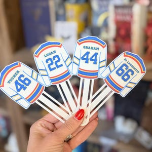 May include: Four white bookmarks shaped like hockey jerseys with blue and yellow trim. Each jersey features a player name and number: Tucker 46, Logan 22, Graham 44, and Di Laurentis 66. The bookmarks have long, flat white stems extending downward.