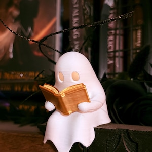 May include: A white ghost figure reading a golden book. The ghost is seated on a dark, textured surface, with a blurred background of books and decorations. The figure has two eye holes and is draped in a sheet.