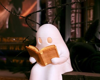Puede incluir: Una figura de fantasma blanco leyendo un libro dorado. El fantasma está sentado sobre una superficie oscura y texturizada, con un fondo borroso de libros y decoraciones. La figura tiene dos agujeros para los ojos y está cubierta con una sábana.